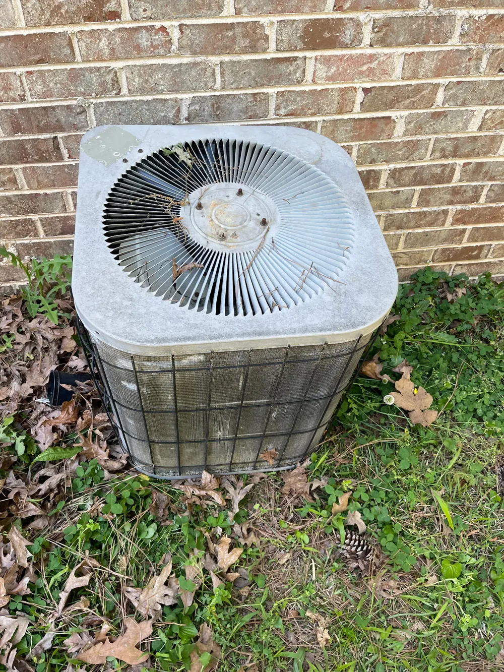 Outdoor AC condenser clogged with leaves and debris beside a brick wall