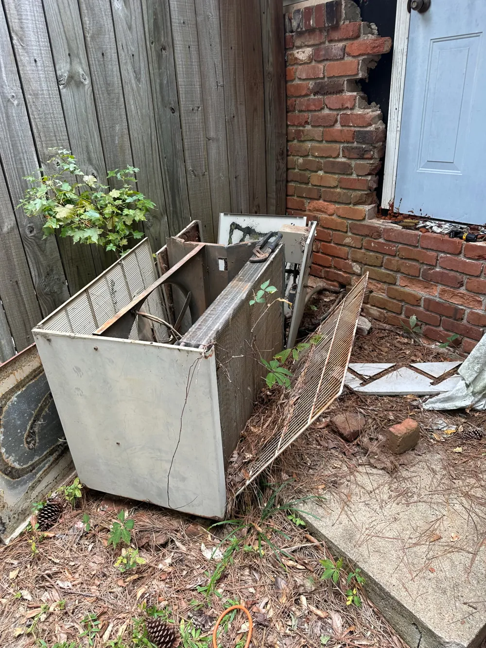 Old, damaged air conditioner cabinet removed and left beside a fence and brick wall